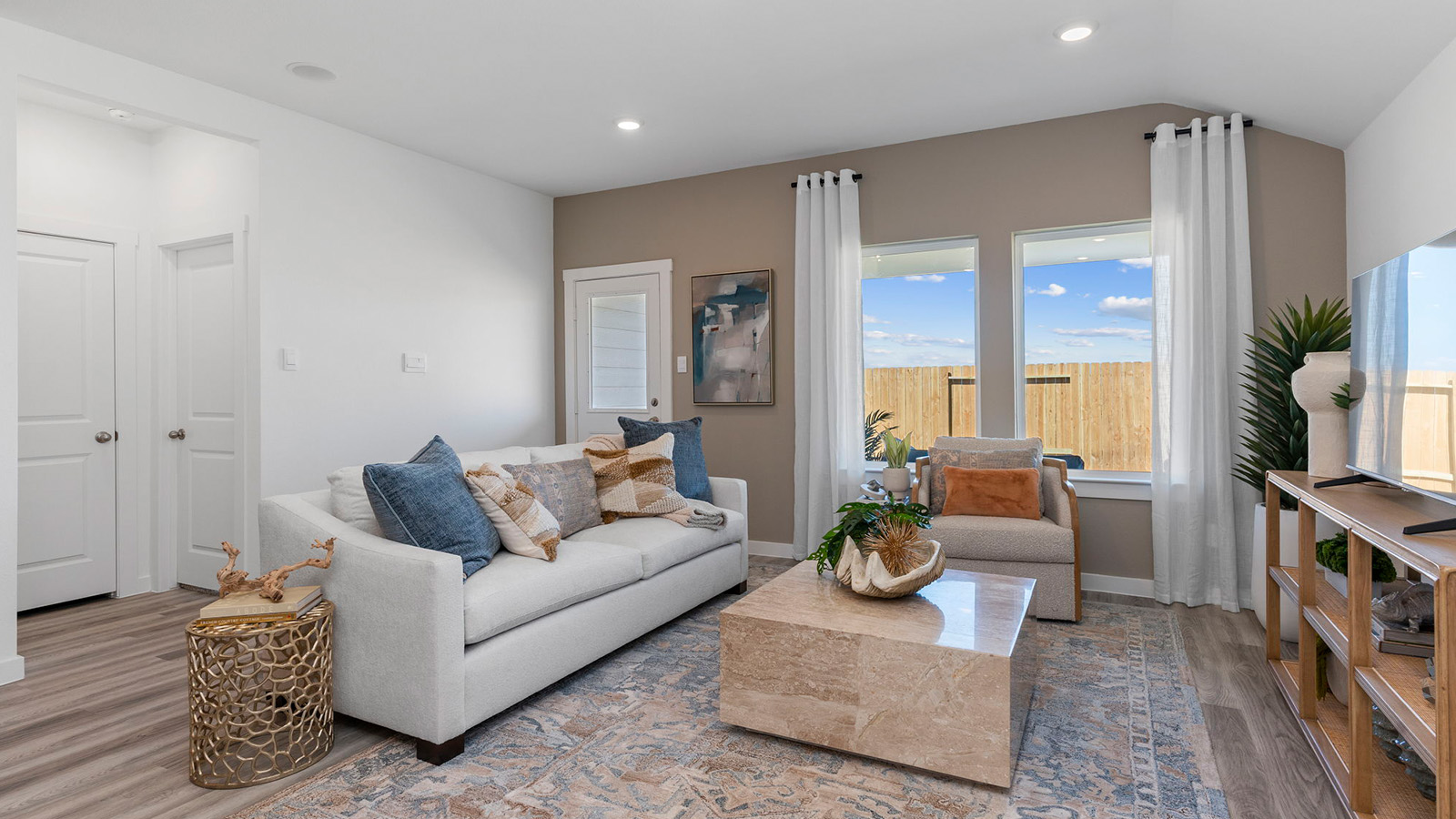 A cozy living room featuring large windows for natural lighting, wood flooring and a view to the kitchen and dining area.