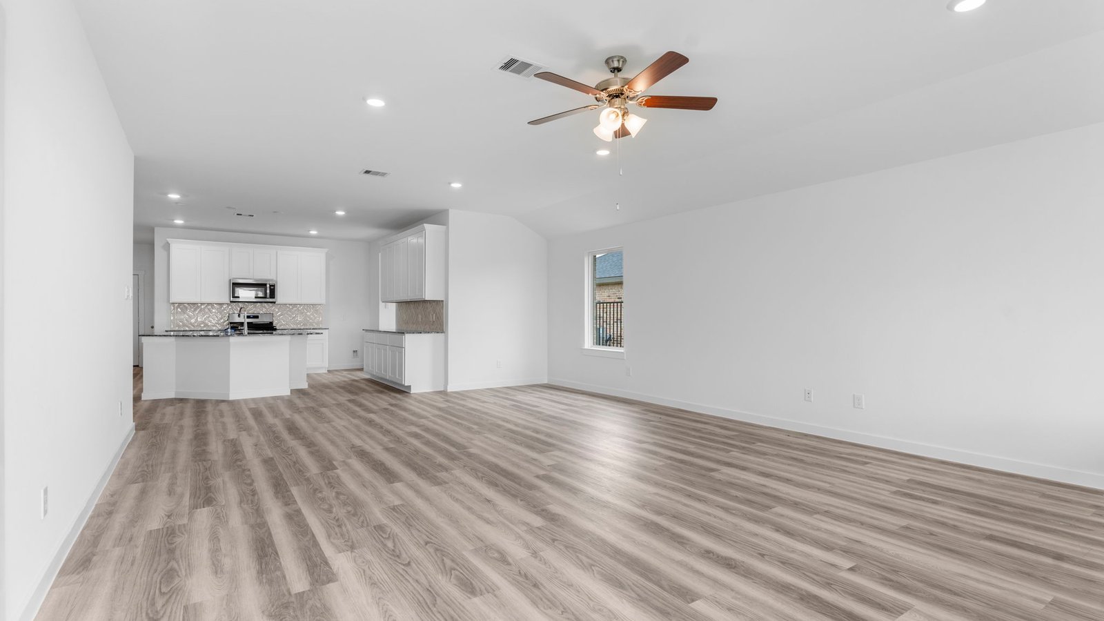 A cozy living room featuring large windows for natural lighting, vinyl wood flooring and a view to the kitchen and dining area.
