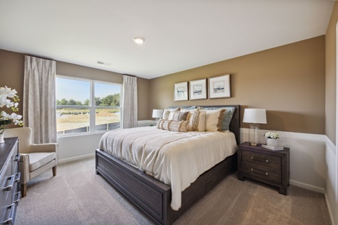 Bedroom with tan walls, beige carpet, large window with white curtains, and bed. Brown bedside table.