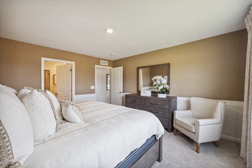 Bedroom with bed, off white chair next to dark brown dresser with mirror, beige carpet and tan walls