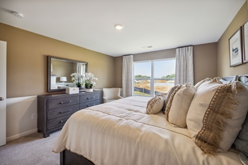 Bedroom with bed, window with white curtains, dark brown dresser with mirror. Tan walls and beige carpet.