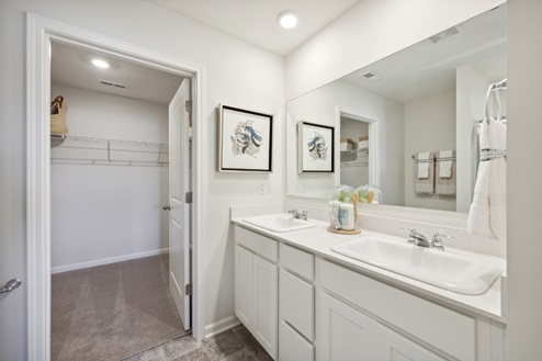 Bathroom with white dual vanity, large mirror above, open door to walk in closet with white wire shelving and beige carpet