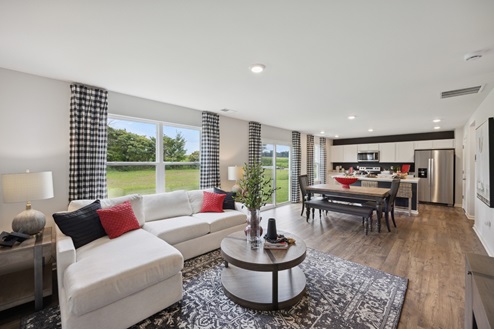 View of open concept living space. White sofa with red and black pillows. Round brown coffee table over black and white area rug. Large windows with black and white curtains. Casual dining table with bench and chairs. White kitchen cabinetry with stainless appliances.