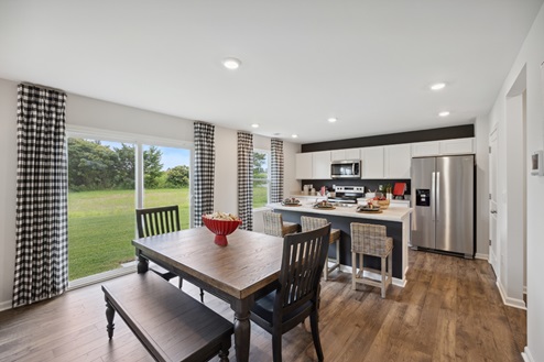 Casual dining area with table with bench and chairs and kitchen with white cabinetry, stainless appliances, white and black island with 3 barstools. Large windows along left wall with black and white patterned curtains. Brown vinyl plank floors and white walls.