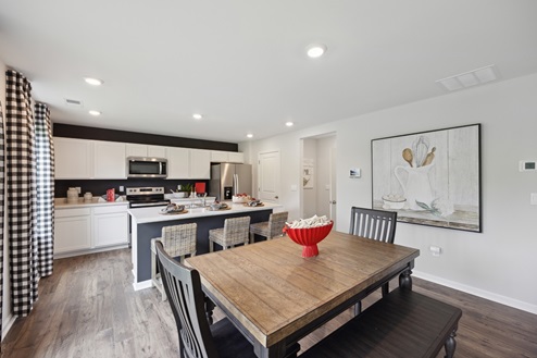 Casual dining table with chairs and bench. Red bowl as centerpiece. Kitchen with white cabinetry and black accent wall. Stainless appliances. Brown vinyl plank floors, white walls with white artwork on right wall and black and white curtains on left wall.