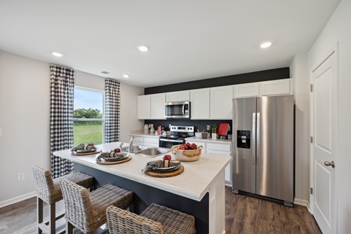 Kitchen with white cabinetry, stainless steel appliances with refrigerator, white island with barstools, window with black and white curtains, white walls, brown vinyl plank floors