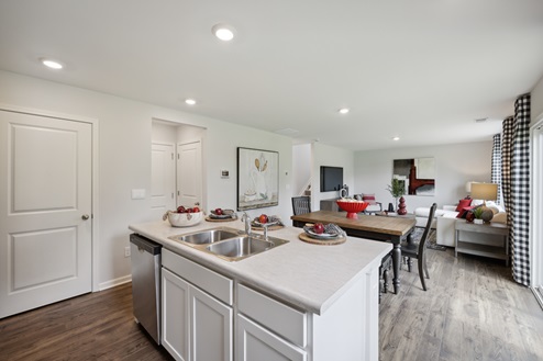 View of open concept living space from kitchen. White island with stainless sink. Casual dining table with dark brown chairs. Living room with white sofa and artwork on white walls. Brown vinyl plank floors, white and black check patterned curtains on left wall.