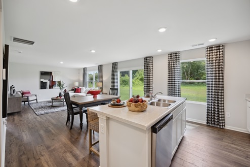 View of open concept living area from kitchen. White island with stainless sink. Casual dining table with chairs. View of living room with furniture. Large windows with black and white check patterned curtains along right wall. Brown vinyl plank floors and white walls.