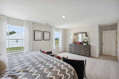 Primary bedroom with bed and 2 chairs and small table in front of bed. Windows along right wall with sheer white curtains. Grey dresser with mirror. Beige carpet and white walls.