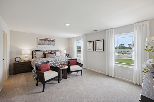 Primary bedroom with bed and 2 chairs and small table in front of bed. Red and black artwork above bed. Brown bedside tables with white lamps. Windows with sheer white curtains along right wall. Beige carpet and white walls.