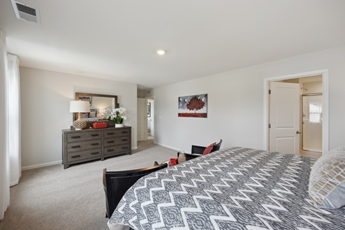 View of primary bedroom from bed with black and white bedding. White walls with red and black artwork on right wall. Open door to en-suite bathroom on right wall. Sheer white curtains along left wall. Brown dresser with mirror. Beige carpet.