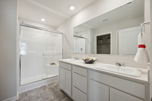 Primary bathroom with white dual vanity and large mirror above. White walk-in shower with glass doors. Tile patterned vinyl floors and white walls.