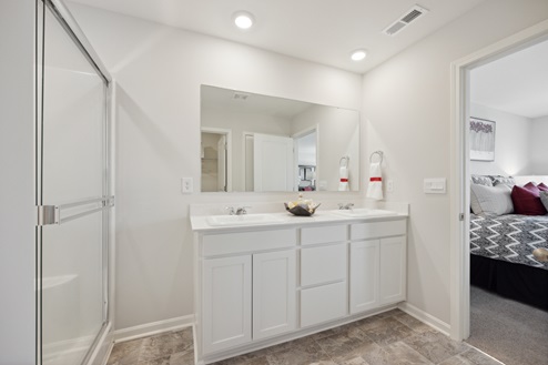 Primary bathroom with white dual vanity and large mirror above. Glass door of walk-in shower to the left. Open door to connected bedroom to the right. White walls, tile patterned vinyl floors.