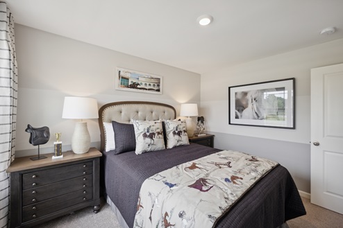 Bedroom with upholstered bed and purple bedding. Brown bedside tables with white lamps. Horse artwork on right wall. Beige carpet and grey and beige striped walls.
