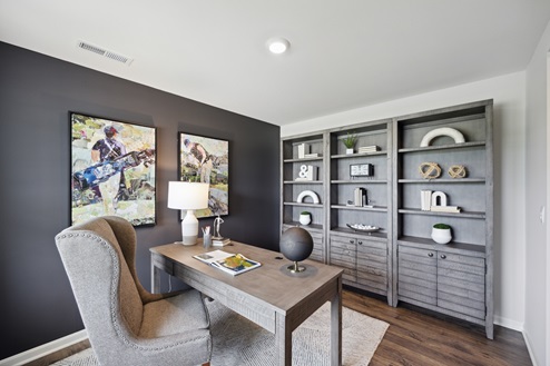 Home office with black accent wall and golf themed artwork. Large grey shelving unit with decor. Light brown desk with chair over off-white area rug on brown vinyl plank floors.