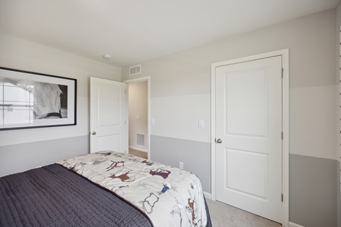 Bedroom with bed with purple bedding and horse patterned throw at the foot of the bed. Lightly painted striped walls with black and white horse artwork. Beige carpet. Closed white closet door and open entry door.
