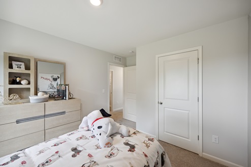 Bedroom with bed with white and dog patterned bedding, light brown dresser to the left. Light grey walls, beige carpet. Closed white closet door. Open entry door.