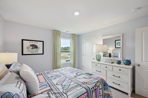 Bedroom with bed with multi-colored bedding, grey walls. White dresser with green lamp and mirror above. Window with green curtains.