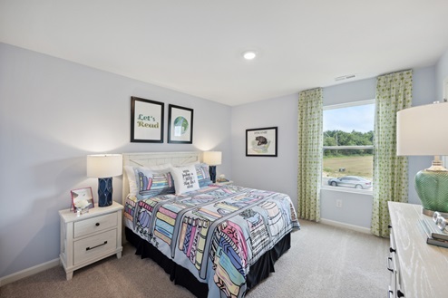 Bedroom with bed with multi-colored bedding, artwork above bed, grey walls and beige carpet. Window with green curtains, white bedside tables with blue lamps, white dresser with green lamp.