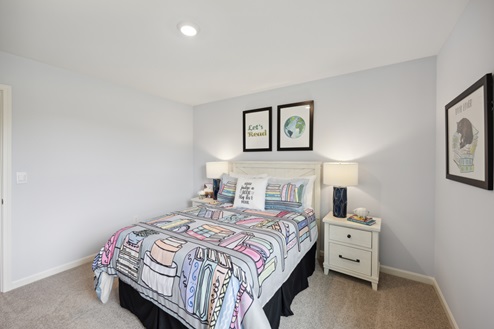 Bedroom with bed and multi-colored bedding, artwork above bed. White bedside tables with dark blue lamps. Beige carpet, grey walls.