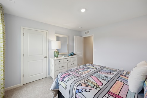 Bed with multi-colored bedding, white dresser with green lamp and mirror, green curtains on window, grey walls, beige carpet. Closed white closet door and open entry door.