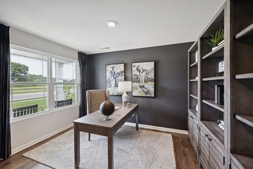 Home office with black accent wall with golf themed artwork. Large windows on the left wall, large grey shelving unit with decor to the right. Light brown desk with chair in center of room over cream and black area rug. Brown vinyl plank floors.