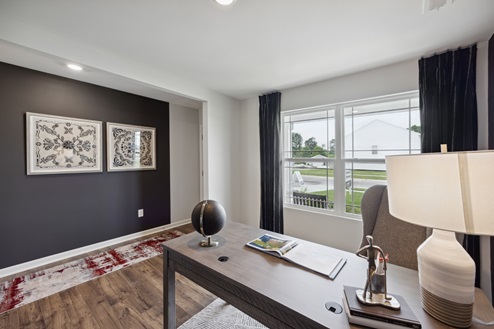 Home office with windows and black curtains, brown desk and chair with white lamp. View into home entry with black accent wall and black and white artwork. Red and cream-colored entry rug over brown vinyl plank floors.