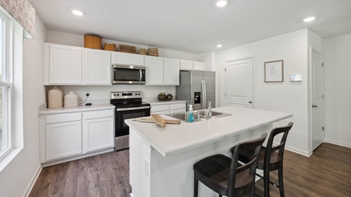 Kitchen with white cabinetry and stainless-steel appliances. Brown baskets above cabinetry. Closed white pantry door to the right. Window with floral valance to the left. White kitchen island with stainless sink. 2 black barstools White walls, brown vinyl plank floors.