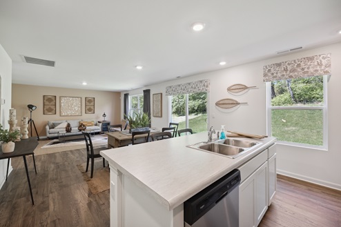 View of open concept living area from white kitchen island with stainless sink and dishwasher. Large windows with floral valances to the right. Light brown dining table with dark brown chairs. Grey sofa with artwork above against tan accent wall.