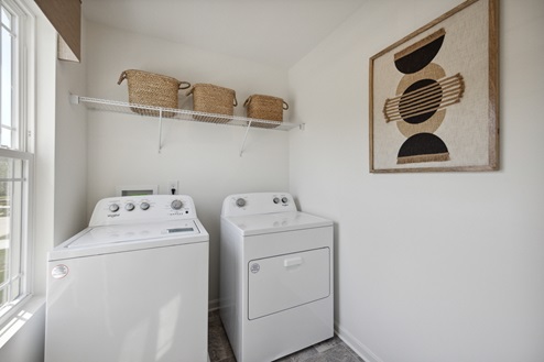 Laundry room with white walls. Window on left wall. White washer and dryer with white wire shelving above. Baskets on shelving. Artwork on right wall.