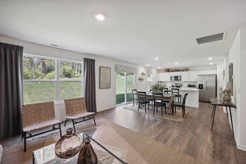Open concept living space. 2 brown chairs against large windows with dark brown curtains. White walls and brown vinyl plank floors. Brown casual dining table with black chairs. Large patio door windows with valance above across from dining table. Kitchen with white cabinetry and stainless appliances.
