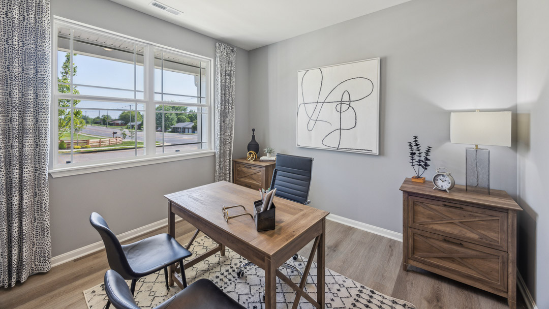 Flex room decorated as a home office with table, chair, 2 client chairs, and accent table with lamp. Double window overlooking the front yard.