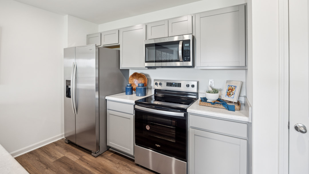 Modern kitchen with stainless steel appliances, gray cabinets, and wood floors. Decor includes a cutting board, blue jars, and a framed picture.