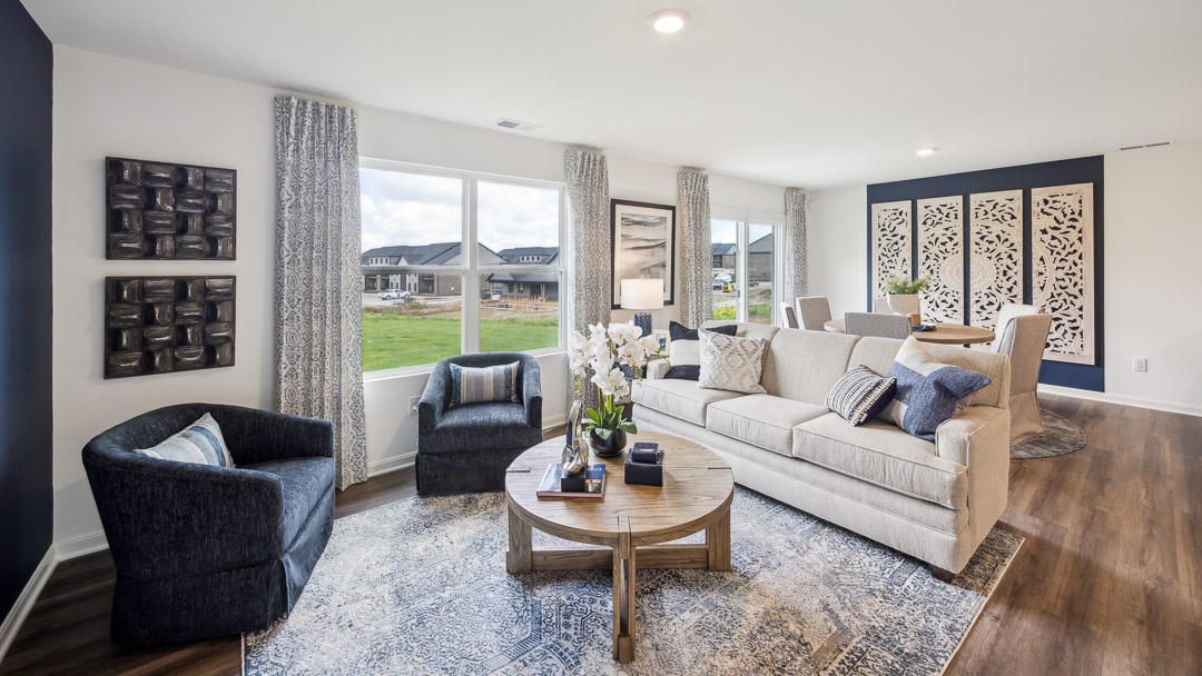 Modern living room with a beige sofa, blue armchairs, and a round wooden coffee table on a patterned rug. Large windows provide natural light, enhancing the neutral tones and cozy ambiance.