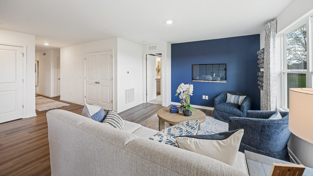 Modern living room with light gray sofa, blue accent wall, round wooden table, and blue chairs. Large window lets in natural light; decor is cozy.