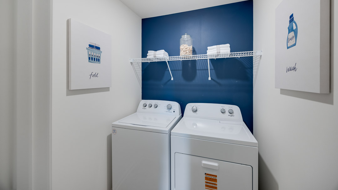 Laundry room with a washer and dryer against a blue wall, featuring a shelf with folded towels and a jar of clothespins. Art prints read "Fold" and "Wash".
