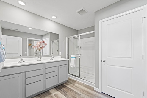 Timber Creek Chatham Model Home - Bathroom off of main bedroom with LVP flooring, walk in shower, and dual vanity sinks with grey cabinetry.