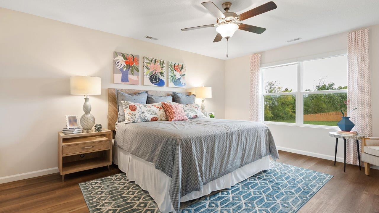 farmstead at carroll creek henley model home primary bedroom with mohawk lvp flooring, 2 windows, ceiling fan, heron plume painted walls, and ensuite walk in closet and bathroom