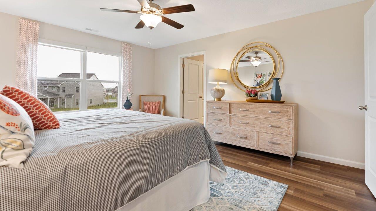 farmstead at carroll creek henley model home primary bedroom with mohawk lvp flooring, 2 windows, ceiling fan, heron plume painted walls, and ensuite walk in closet and bathroom