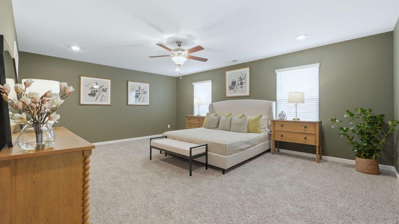 thunderhawk model home elm plan primary bedroom with beige carpet flooring, sage green walls, 2 large windows, with space for seating and ensuite bath and closet