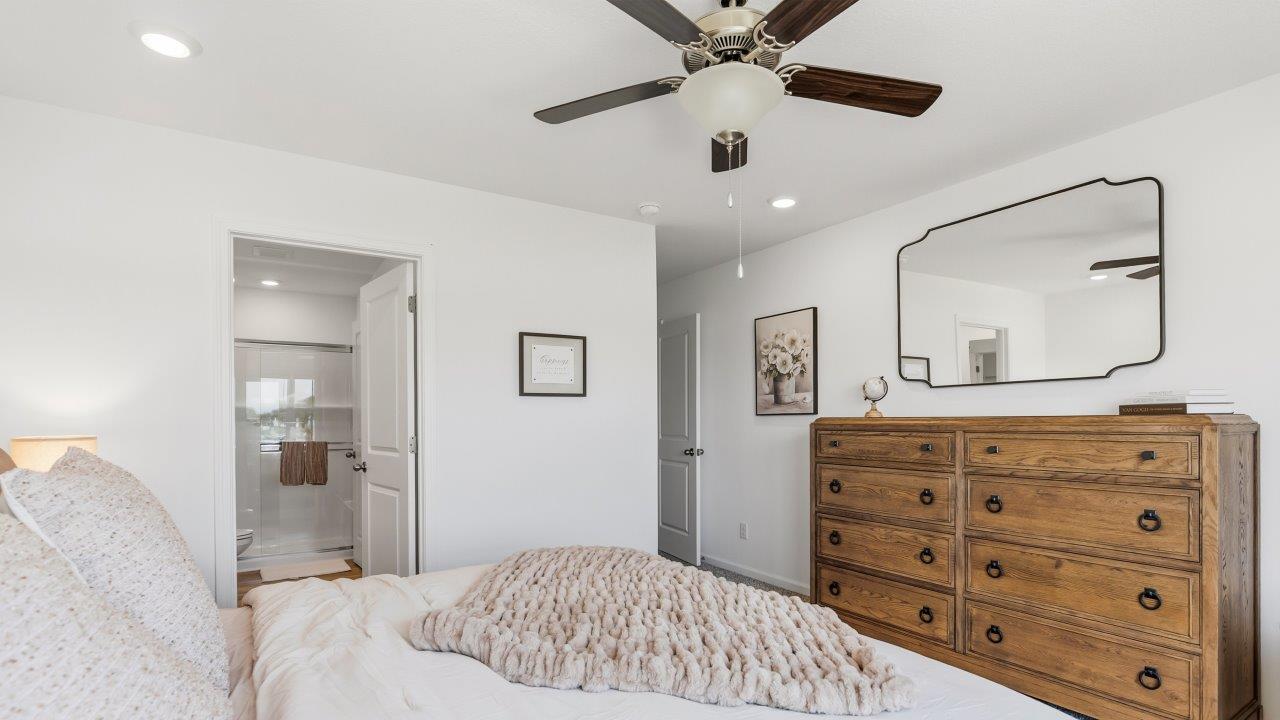 Whispering Pines Pattison Model - primary bedroom with carpet flooring, ceiling fan, white walls, 2 windows, and ensuite bathroom and walk in closet