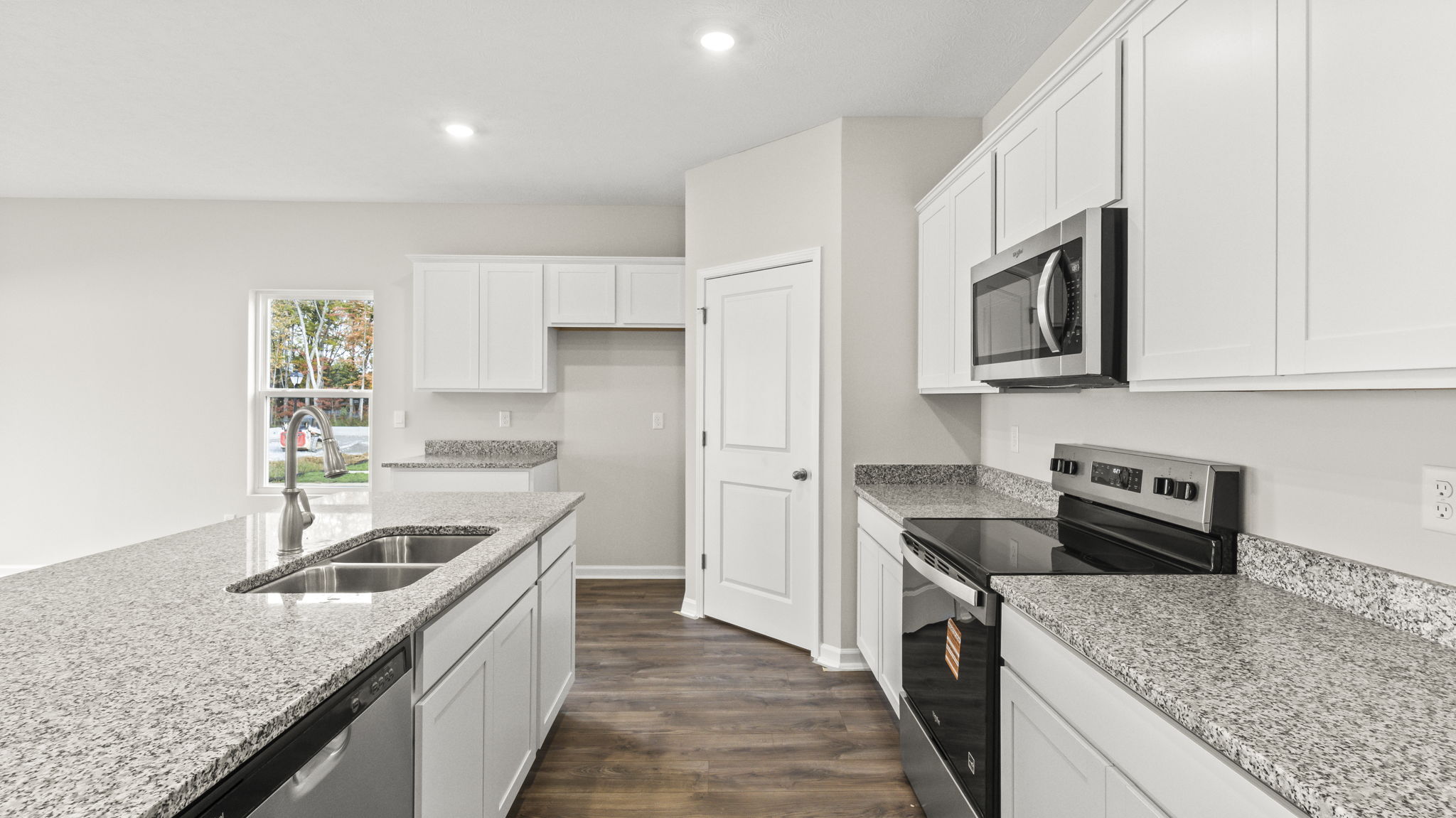 Side view of kitchen with granite countertops, island with build-in dishwasher on the left, appliances on the right, and door to walk-in pantry