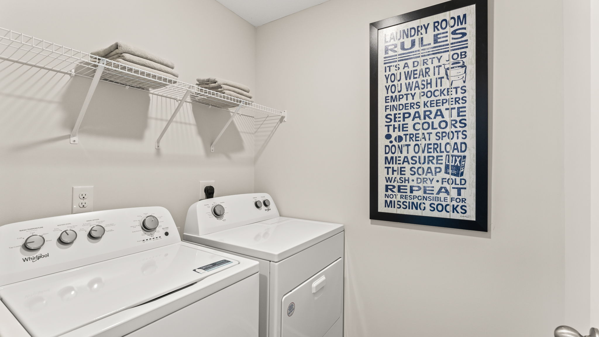 Closeup of washer/dryer with wire rack shelving above and art on the right.
