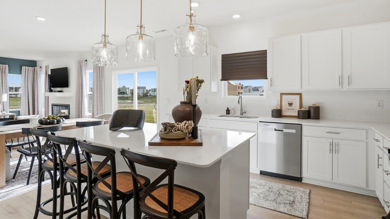 Galley view through a white kitchen with a large island, double sink, pendant lights, dining area, and living room beyond.