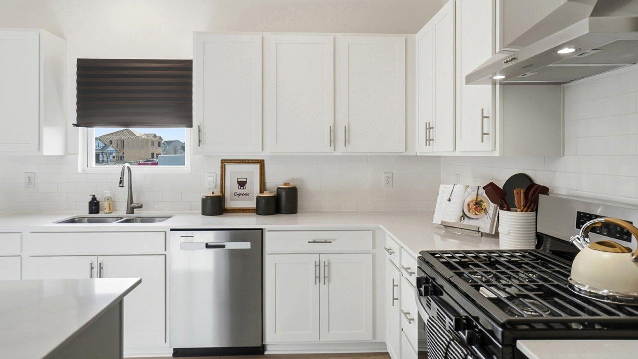 White kitchen island with four black stools, glass pendant lights, stainless steel dishwasher, and living area visible beyond.