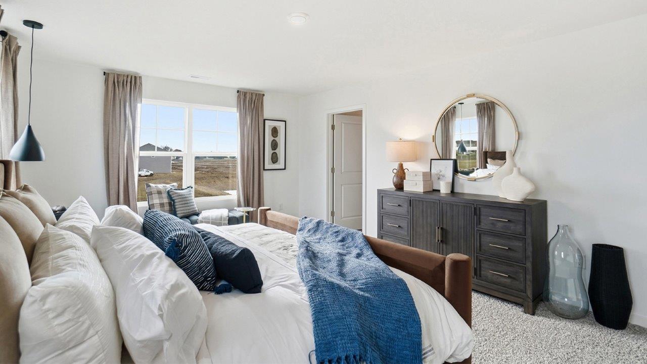 Primary bedroom viewed across a brown upholstered bed toward a dark dresser, round mirror, black-and-white artwork, and adjoining bathroom.
