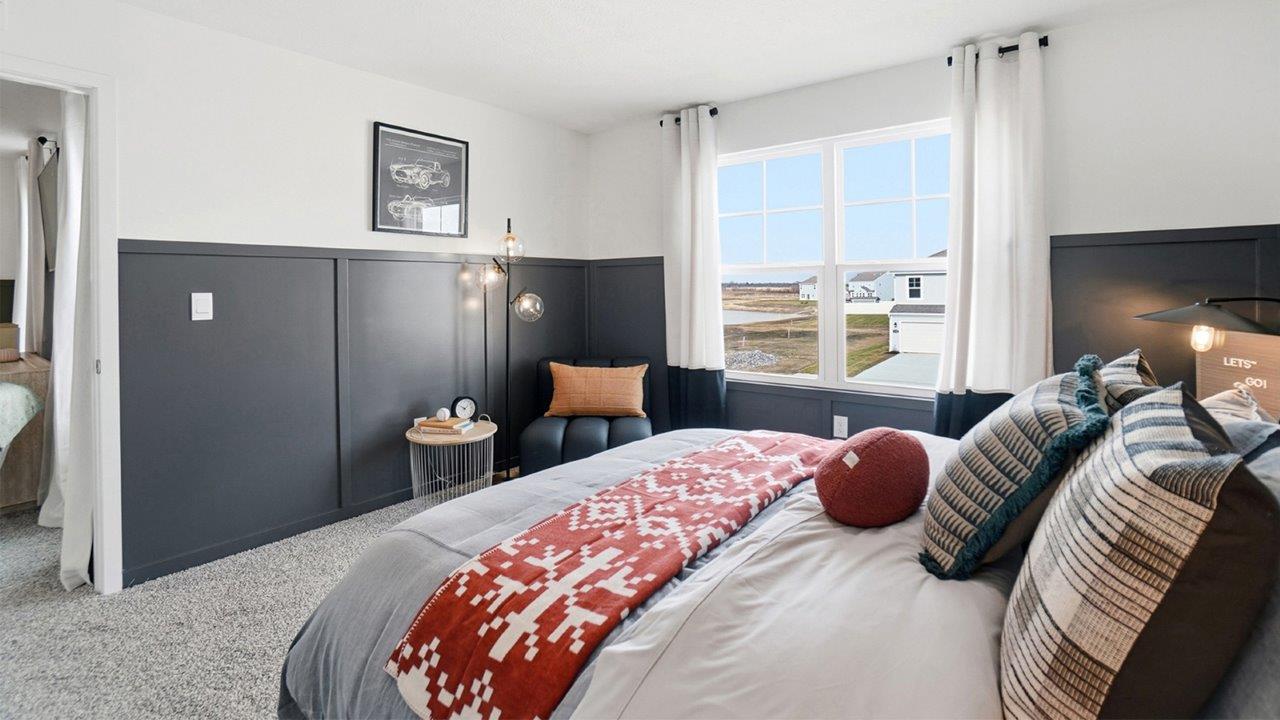 Bedroom viewed across a gray bed toward a sunny window, with dark wall paneling and a small seating area.