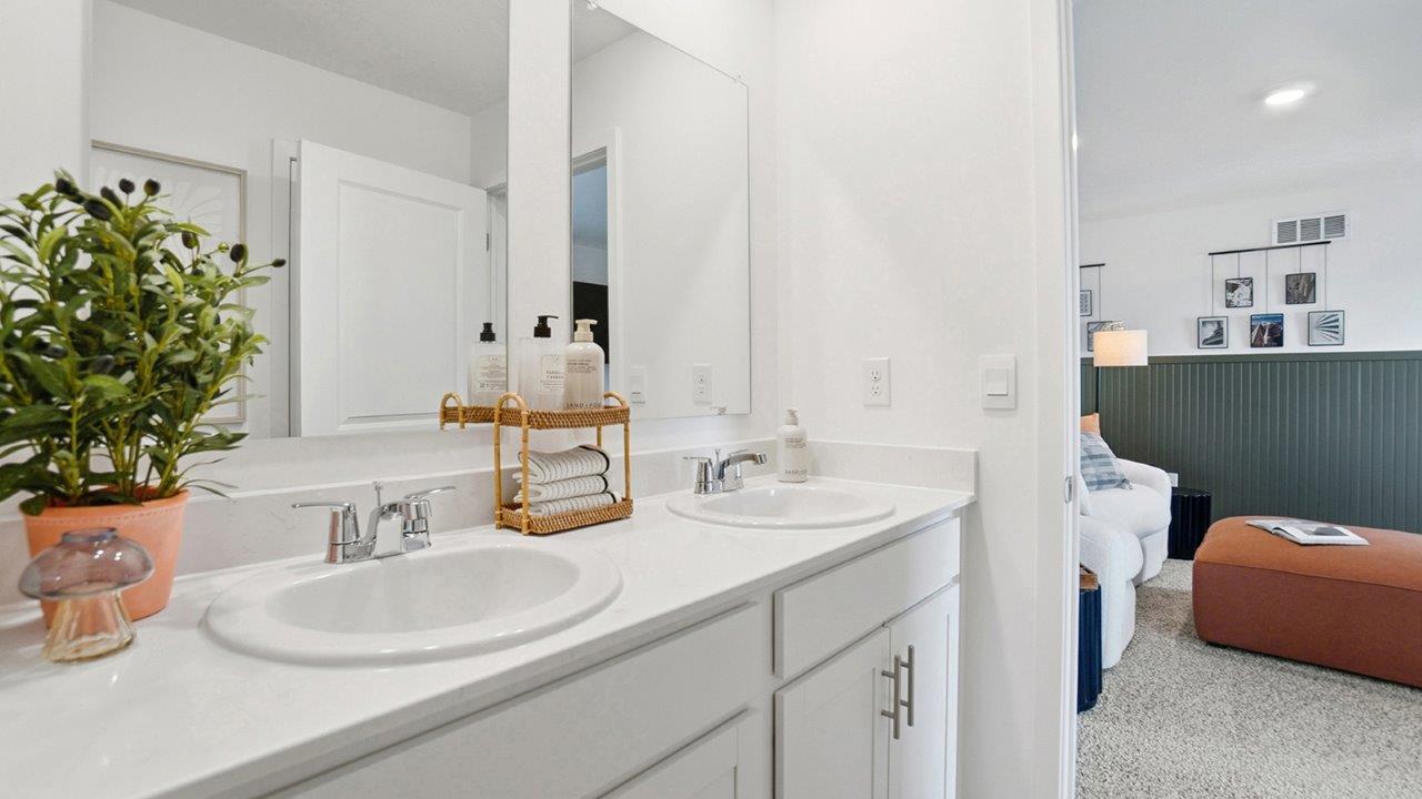 Double-sink white vanity with large mirrors and greenery, opening into a carpeted bedroom with an orange bench.
