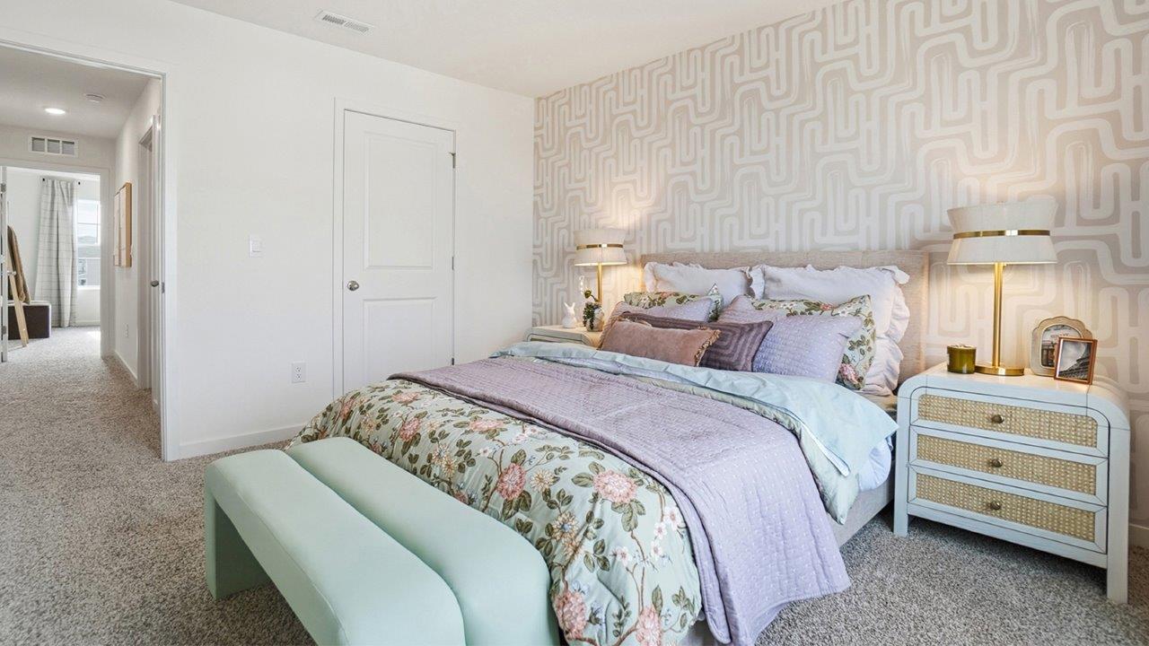 Bedroom with floral bedding, pale green bench, woven-front nightstands, and a soft geometric accent wall.