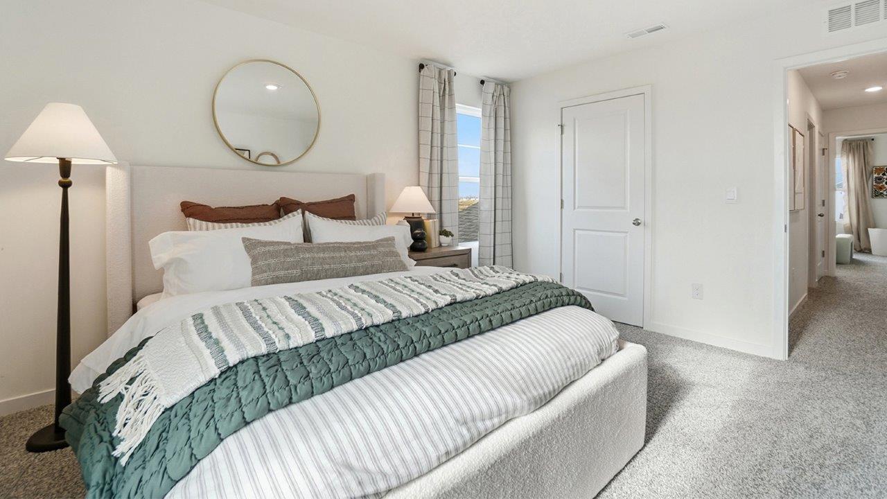 White upholstered bed with layered neutral bedding and green throw, beside lamps, a round mirror, and hallway entrance.